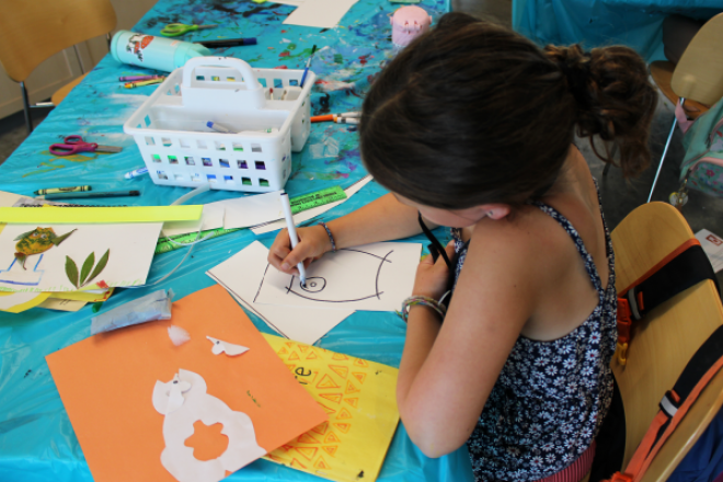 A child draws with marker at a table covered in various art supplies.