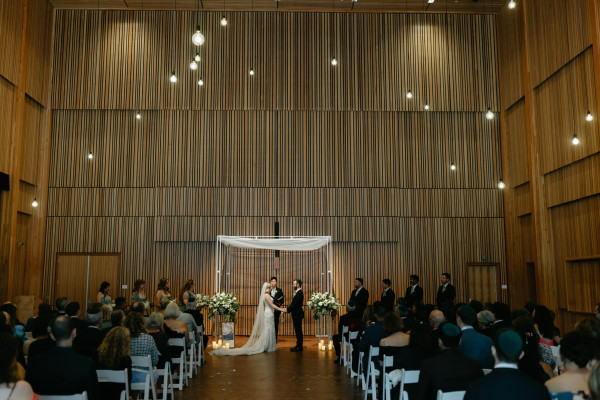 Two people hold hands during their marriage ceremony in Osberg Great Hall. Pendant lights hang from the ceiling and contrast against the wood-clad walls of the Hall.