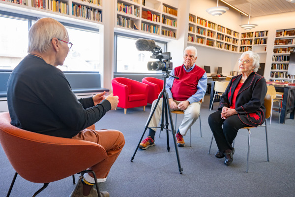 A person sits in front of a film camera in the Library, which is brightly lit and has floor-to-ceiling bookshelves lining many of the walls.
